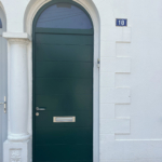 Porte d’entrée en bois vert avec imposte vitrée en plein cintre et entrée de courrier inox, sur une façade blanche à Biarritz.