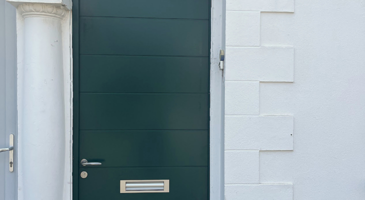 Porte d’entrée en bois vert avec imposte vitrée en plein cintre et entrée de courrier inox, sur une façade blanche à Biarritz.