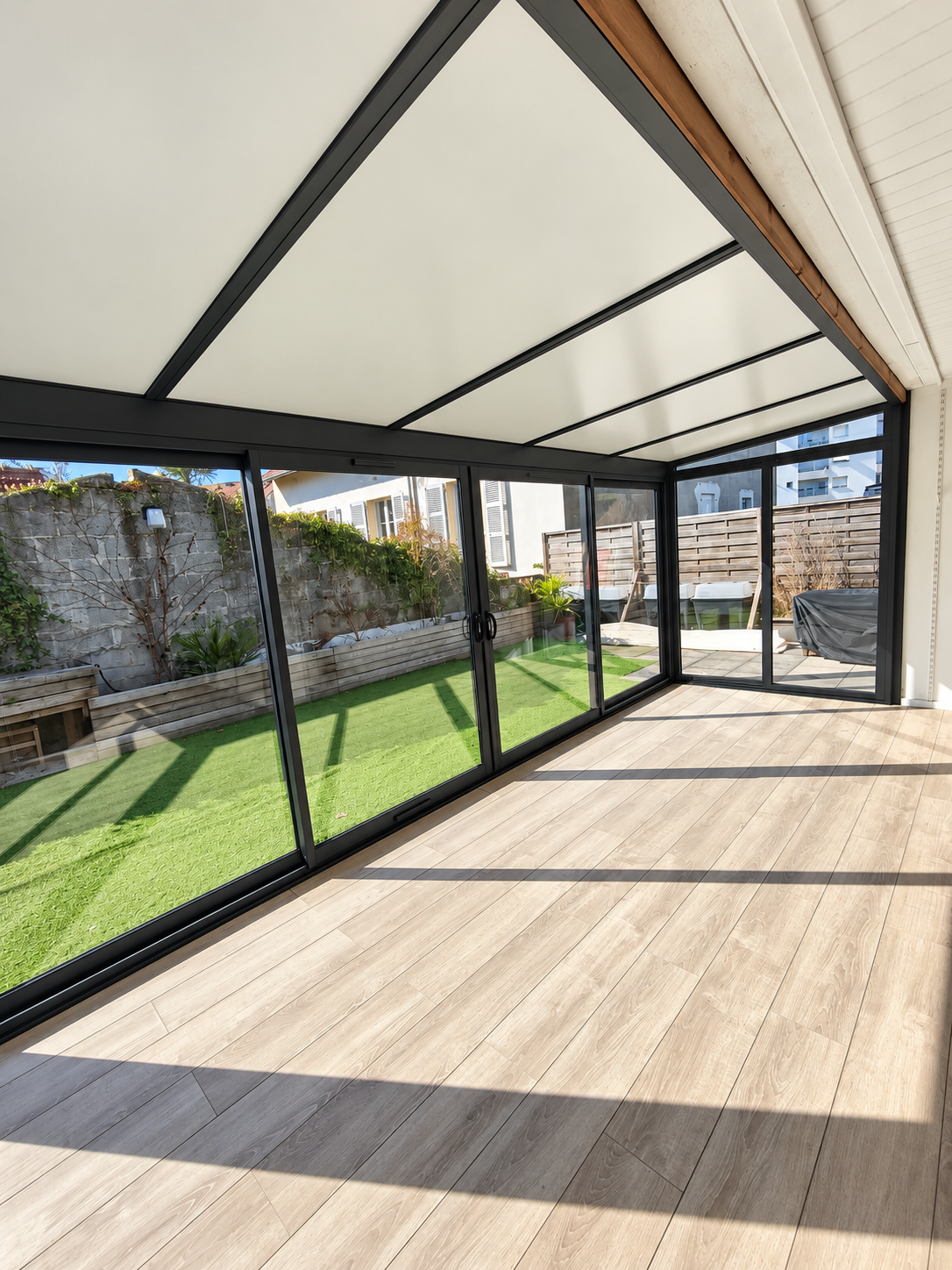 Intérieur de la véranda rénovée, avec structure noire, toiture claire et vue sur le jardin